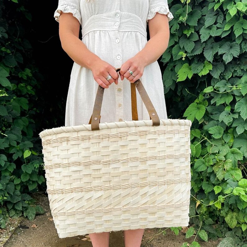 May include: A cream-coloured, rectangular woven basket with brown leather handles. The basket has a textured pattern. The person holding the basket is wearing a light-coloured dress. The background features green foliage.
