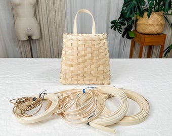 May include: A light-colored, woven basket with a handle, standing upright. Several bundles of thin, light-colored, woven materials are in front of the basket. The basket and materials appear to be for crafting.