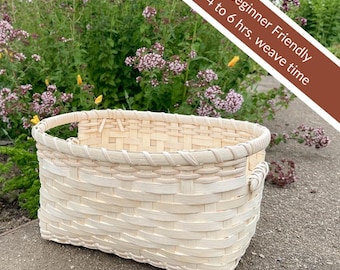 May include: A rectangular, light-colored woven basket with a handle. The basket is made of natural-colored woven material. The text "Beginner Friendly 4 to 6 hrs. weave time" is in the upper right corner. The background features green plants and purple flowers.