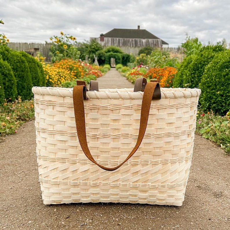 May include: A rectangular, light-coloured woven basket with brown leather handles. The basket is made of woven strips of wood, and the handles are attached to the top. The basket is on a paved path, with a garden and building in the background.