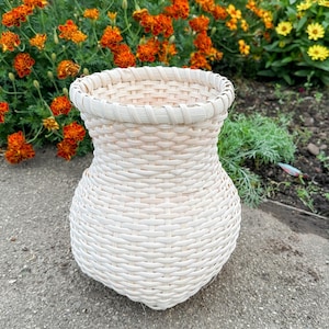 May include: A handwoven, off-white basket with a rounded base and a flared rim. The basket is made of woven natural fibers and sits on a concrete surface, with a backdrop of orange and yellow flowers.