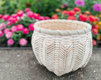 May include: A round, woven basket with a light beige color. The basket has a textured, chevron-like pattern on the sides and a braided rim. The background features a blurred garden with pink, red, and yellow flowers.