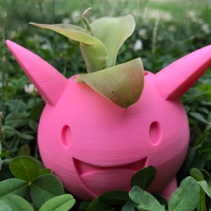 May include: A pink 3D printed planter shaped like a cartoon character with a smiling face and two ears. A small green plant is growing inside the planter.