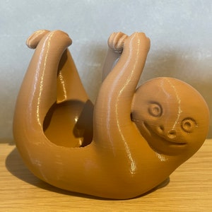 May include: A brown ceramic sloth planter with a smiling face and outstretched arms. The planter is designed to hold small plants or succulents.