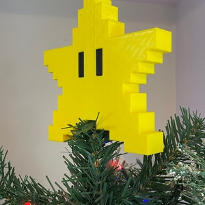 Pode incluir: Uma estrela amarela pixelada com olhos pretos, um personagem popular de videogame, usado como topo de árvore de Natal.