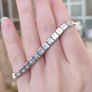 May include: Silver bracelet with square links, each decorated with a tiny star. The bracelet is displayed on a hand, highlighting its design. A simple and elegant piece of jewellery.