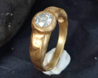 May include: A gold-colored ring with a large, clear, round gemstone. The ring is set on a small, white, irregular-shaped base. The background is dark, with a blurred, textured appearance.