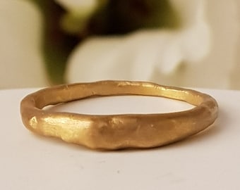 May include: A gold ring with a textured finish. The ring has a simple, organic shape and is likely made of metal. The ring is shown on a white surface with a blurred floral backdrop.