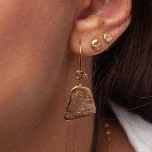 May include: Gold dangle earrings with a textured triangular pendant and a smaller round charm. The earrings are worn in a person's ear.