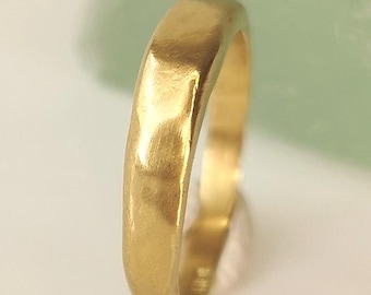 May include: A gold ring with a slightly textured surface. The ring has a rectangular shape and a polished finish. The ring is set against a soft, blurred background.