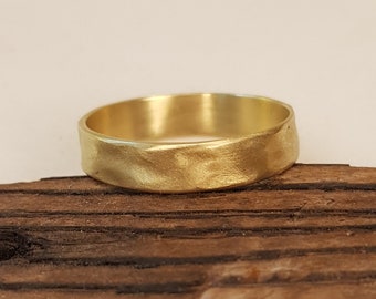 May include: A gold-colored ring with a textured surface. The ring is set against a dark brown wooden background. The ring's band has a slightly uneven texture, giving it a unique, handcrafted appearance.