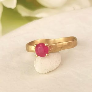 May include: A gold ring with a bright red ruby gemstone set in the center. The ring band is textured and has a hammered finish.