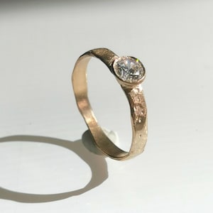 May include: A gold ring with a textured band and a single, clear, round gemstone. The ring is set against a white background, with a shadow cast to the left. The ring's design is simple and elegant.
