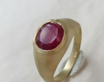 May include: A gold ring featuring a large, oval-cut ruby gemstone. The ruby is a deep red color and is set in a simple, brushed gold band. The ring has a classic, elegant design.