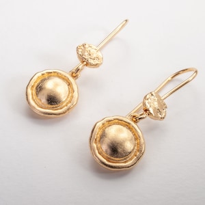 Op de afbeelding: Een paar gouden oorbellen met een ronde, getextureerde schijf en een kleinere, onregelmatig gevormde schijf die aan elke oorbel hangt.