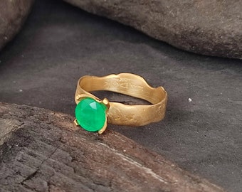 May include: A gold ring with a textured band and a green gemstone. The ring is set on a dark gray background.