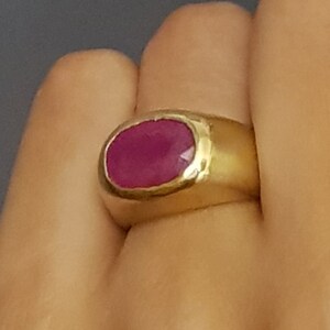 Könnte beinhalten: Ein goldener Ring mit einem großen ovalen Rubin-Edelstein. Der Ring ist in einer schlichten Bandfassung mit leicht strukturierter Oberfläche gefasst.