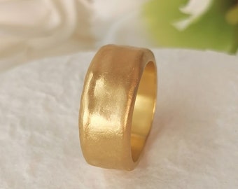 May include: A gold-colored ring with a textured surface. The ring has a wide band and a smooth interior. The ring is likely made of metal and has a matte finish. The ring is a piece of jewelry.