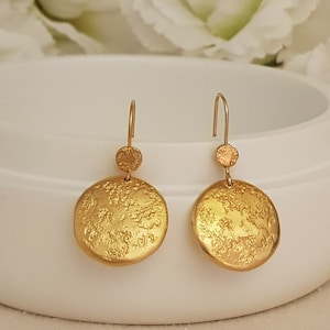 May include: A pair of gold-toned dangle earrings featuring a textured round disc and a smaller round accent. The earrings have a simple, minimalist design.