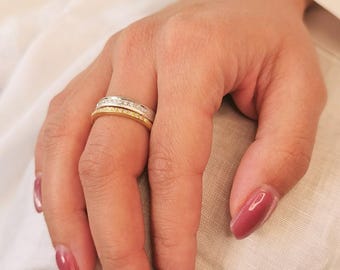 May include: A hand wearing two rings. The top ring is silver with a row of small, clear stones. The bottom ring is gold with a row of small, clear stones. The nails are painted a deep pink color.