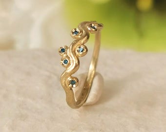 May include: A gold ring with a wavy design, adorned with small, round, blue gemstones. The ring is set against a soft, white background, highlighting its intricate details and craftsmanship. The ring's design is organic and flowing.