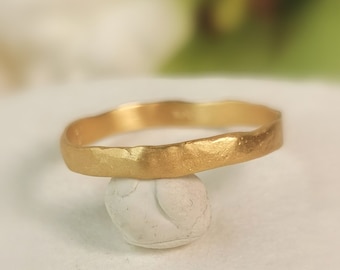 May include: A gold-colored ring with a textured surface. The ring has a wavy design and is displayed on a small, white, rounded object. The ring's design is simple and elegant.