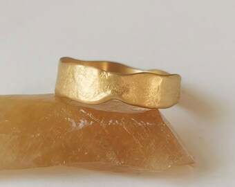 May include: A gold-colored ring with a wavy, textured surface. The ring is sitting on a light brown rock.