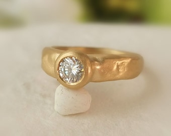 May include: A gold ring with a round, clear diamond set in a bezel setting. The band is textured and has a hammered finish.