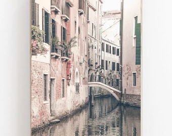 May include: A narrow canal in Venice, Italy, with a small bridge and buildings on either side. The buildings are painted in soft, muted colors, and the water is calm and reflective.