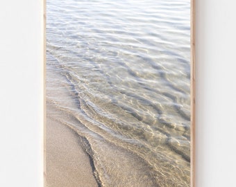 May include: A close-up photograph of clear, shallow water lapping onto a sandy beach. The water is rippling and reflecting the sunlight, creating a shimmering effect.