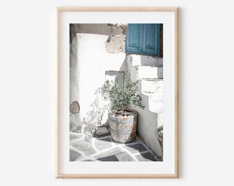 May include: A framed print featuring a sunlit outdoor scene. A small olive tree in a weathered wooden barrel sits on a patterned stone floor. White walls and stairs lead to a blue shuttered window, creating a serene, Mediterranean aesthetic.