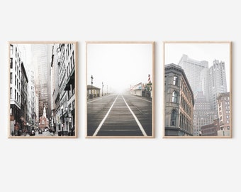 May include: Set of three framed prints featuring urban scenes. The prints depict cityscapes with tall buildings, a boardwalk, and a street scene. The color palette is primarily black, white, and gray, creating a neutral and modern aesthetic.