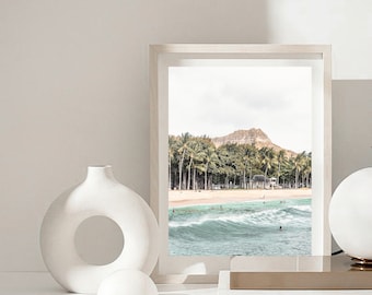 May include: A framed print of a beach scene with palm trees, a mountain, and ocean waves. The print is in a light-colored frame and is displayed with white decorative vases and a lamp.