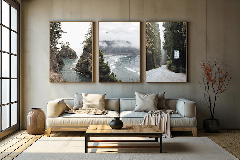 May include: Three framed landscape prints depicting coastal and forest scenes. The artwork features a coastal view with rocky islands, a misty ocean view, and a road through a forest. The prints are displayed above a beige sofa.