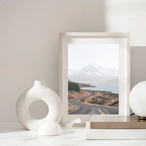 May include: A framed photograph of a winding road leading to a mountain range and a lake. The photo is in a white frame and is sitting on a white surface with a white vase and a white sphere.
