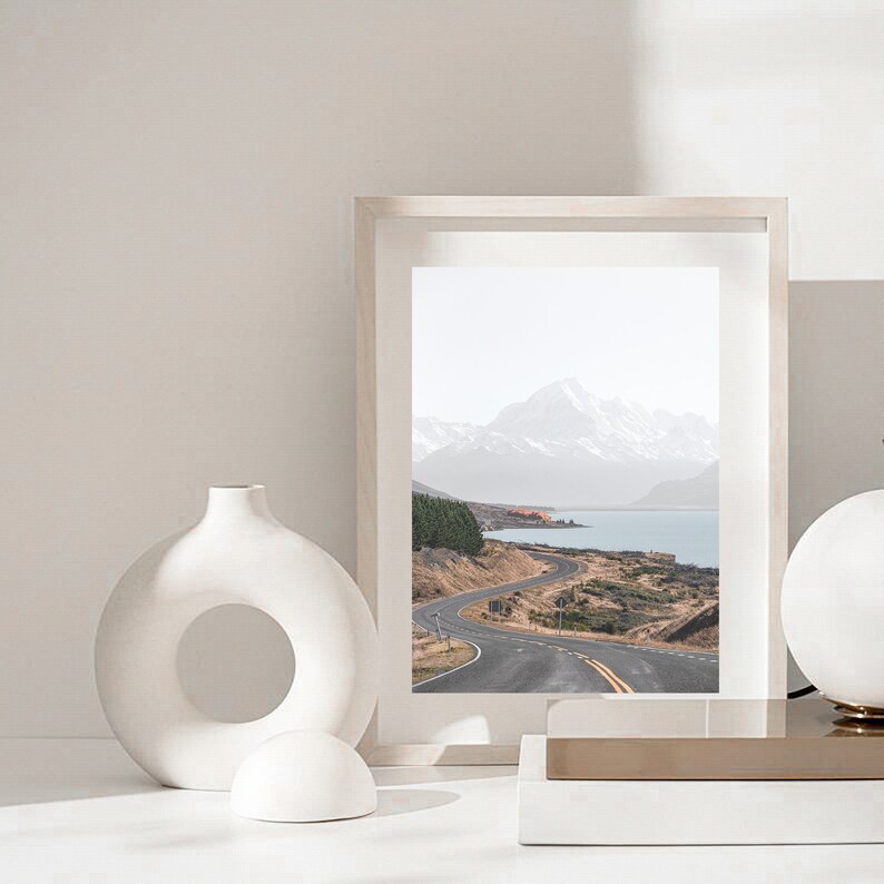 May include: A framed photograph of a winding road leading to a mountain range and a lake. The photo is in a white frame and is sitting on a white surface with a white vase and a white sphere.