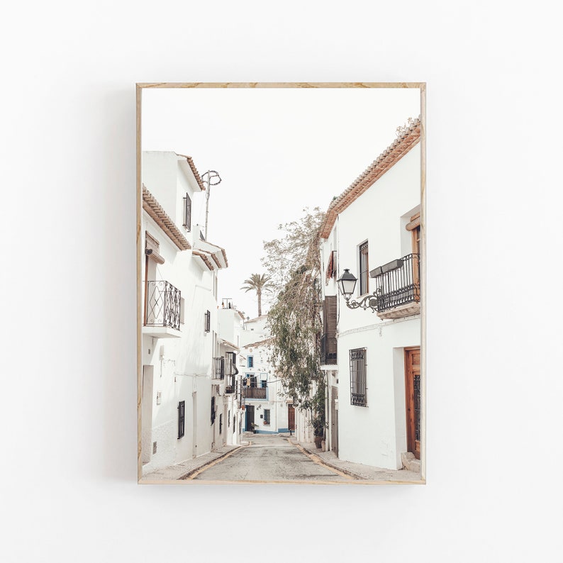 May include: A narrow, white-washed street in a European town with a palm tree in the distance. The street is lined with white buildings with terracotta tile roofs and wrought iron balconies.