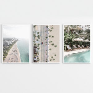 May include: Three framed prints of beach scenes. The first print shows an aerial view of a beach with white sand, blue water, and palm trees. The second print shows a close-up of a beach with palm trees and a red car driving down the road. The third print shows a pool with lounge chairs and palm trees.