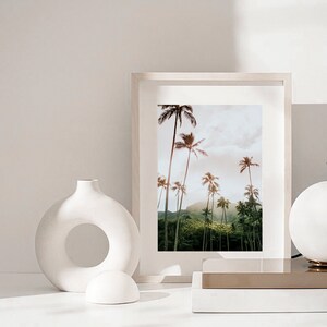 May include: A white framed print of a tropical landscape with palm trees and lush greenery. The print is hanging on a white wall with a white ceramic vase and a white ceramic sphere in the foreground.
