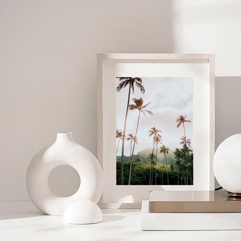 May include: A white framed print of a tropical landscape with palm trees and lush greenery. The print is hanging on a white wall with a white ceramic vase and a white ceramic sphere in the foreground.