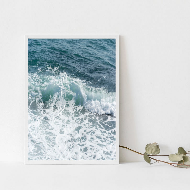 May include: A framed print showing a close-up of ocean waves. The image captures the movement of the water, with shades of teal and white creating depth and texture. The white frame complements the cool tones of the ocean scene, evoking a coastal theme.