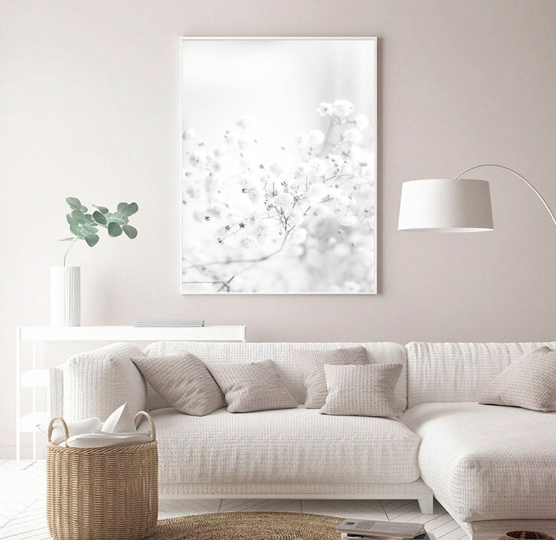 Pode incluir: Uma fotografia a preto e branco de um ramo de flor delicado com pequenas flores brancas. A imagem est&aacute; emoldurada e pendurada numa parede branca acima de um sof&aacute; branco com almofadas.