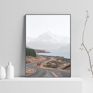 May include: A winding road leads through a mountain landscape with a lake in the distance. The mountains are snow-capped and the sky is cloudy.
