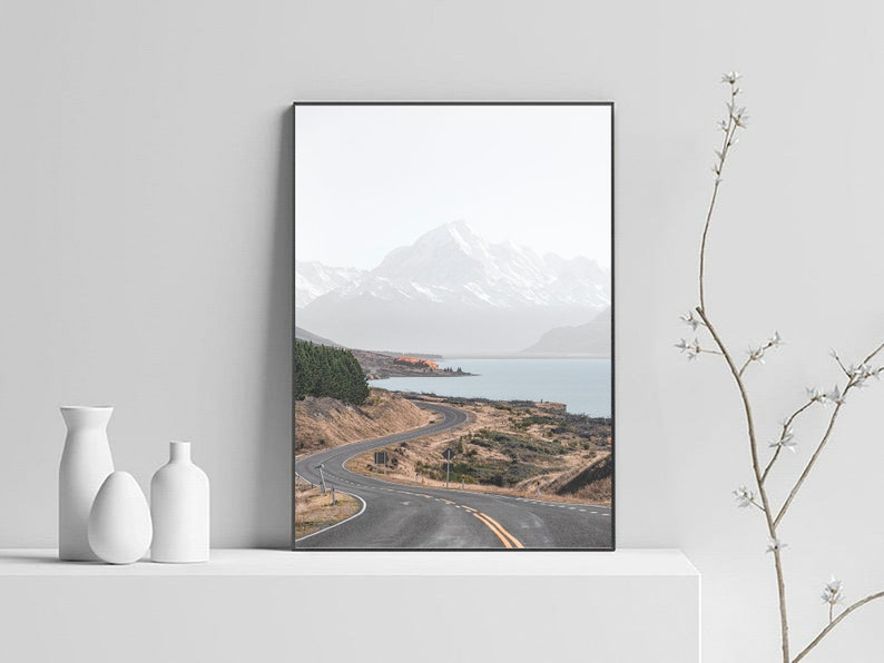 May include: A winding road leads through a mountain landscape with a lake in the distance. The mountains are snow-capped and the sky is cloudy.
