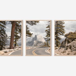 May include: Set of three framed landscape prints featuring a winding road through a mountain range. The prints showcase a scenic view with trees, a road, and a mountain peak under a cloudy sky. The frames are a light wood color.