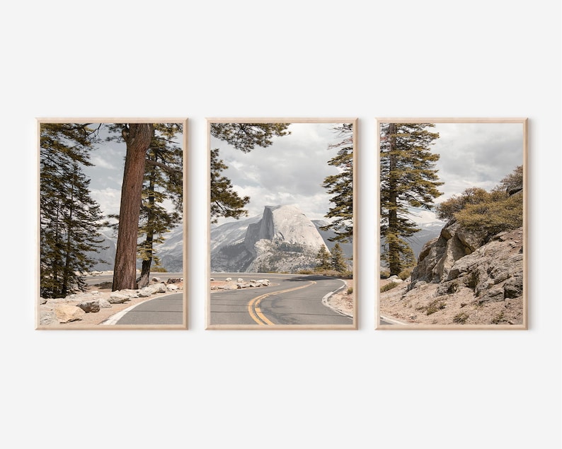 May include: Set of three framed landscape prints featuring a winding road through a mountain range. The prints showcase a scenic view with trees, a road, and a mountain peak under a cloudy sky. The frames are a light wood color.