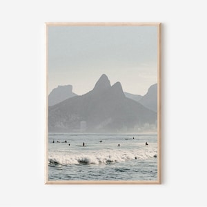 May include: A photograph of a beach with a mountain range in the background. The water is blue and there are waves crashing on the shore. There are several people surfing in the water.