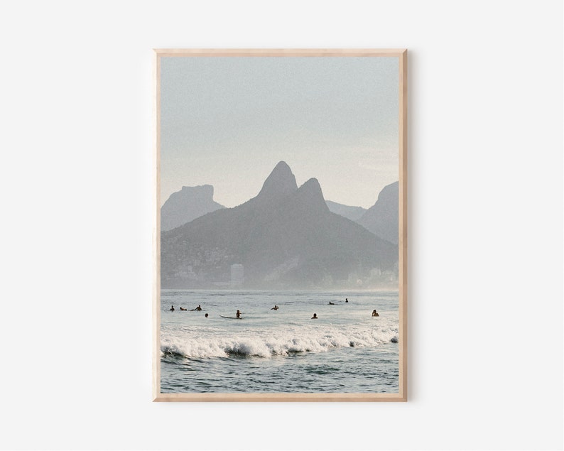 May include: A photograph of a beach with a mountain range in the background. The water is blue and there are waves crashing on the shore. There are several people surfing in the water.