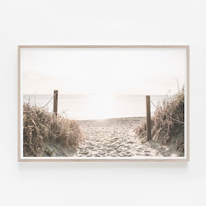 May include: A pathway through tall grass leads to a sandy beach with the ocean in the distance. The sun is shining brightly, creating a warm glow over the scene.