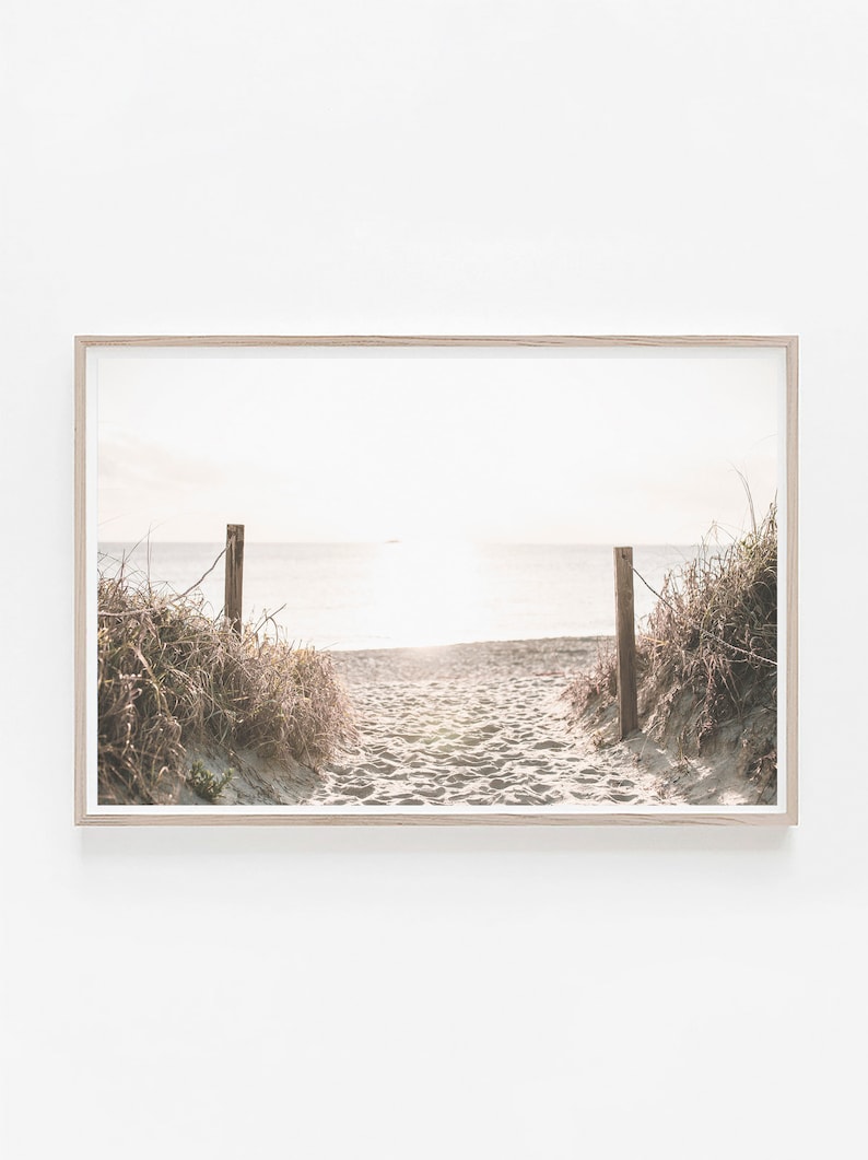 May include: A pathway through tall grass leads to a sandy beach with the ocean in the distance. The sun is shining brightly, creating a warm glow over the scene.